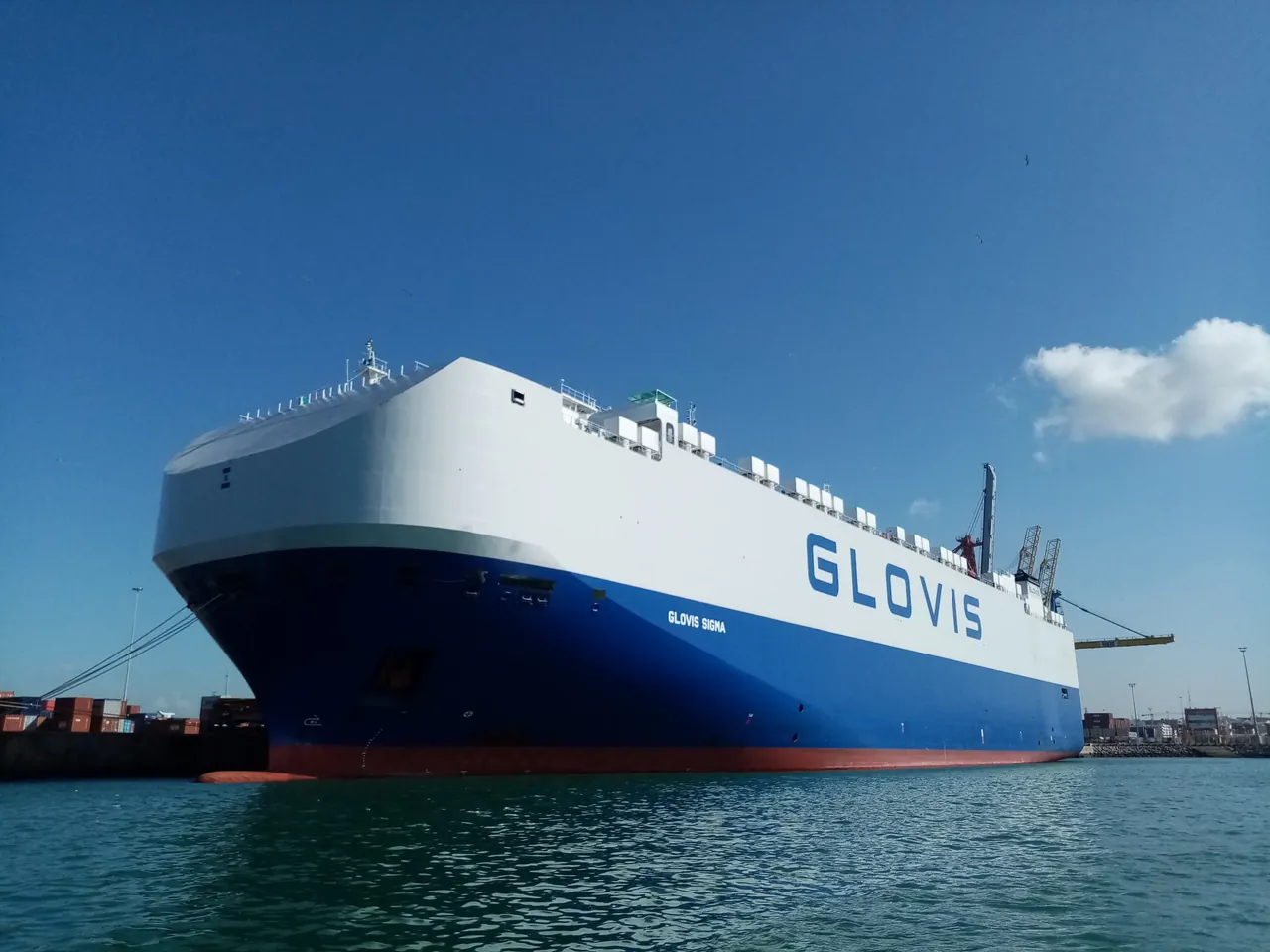Barco portacoches Glovis Sigma, con el casco azul y blanco, atracado en un puerto bajo un cielo despejado